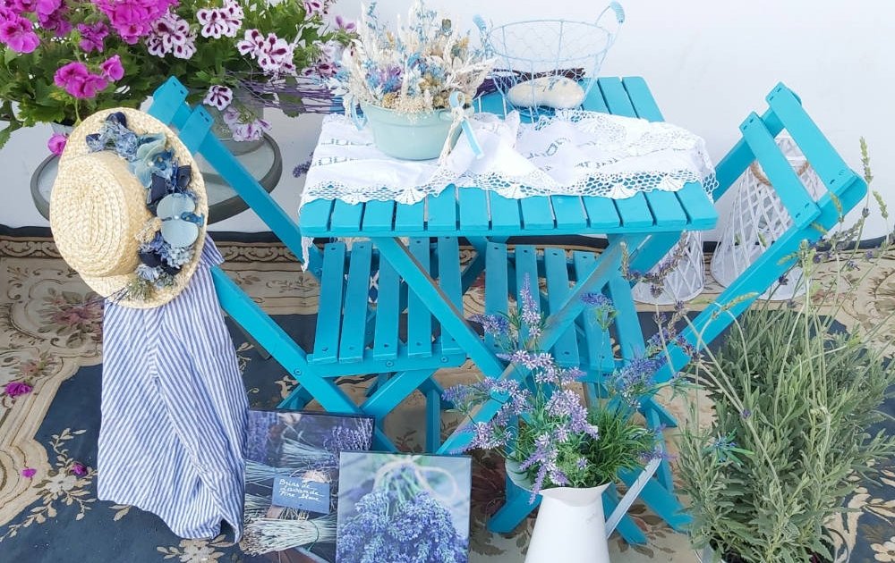 Una terrraza con muebles de madera de color azul, planta lavanda, flores preservadas, sombrero decorativo con flores y lienzos con fotografía de ramilletes de lavanda.