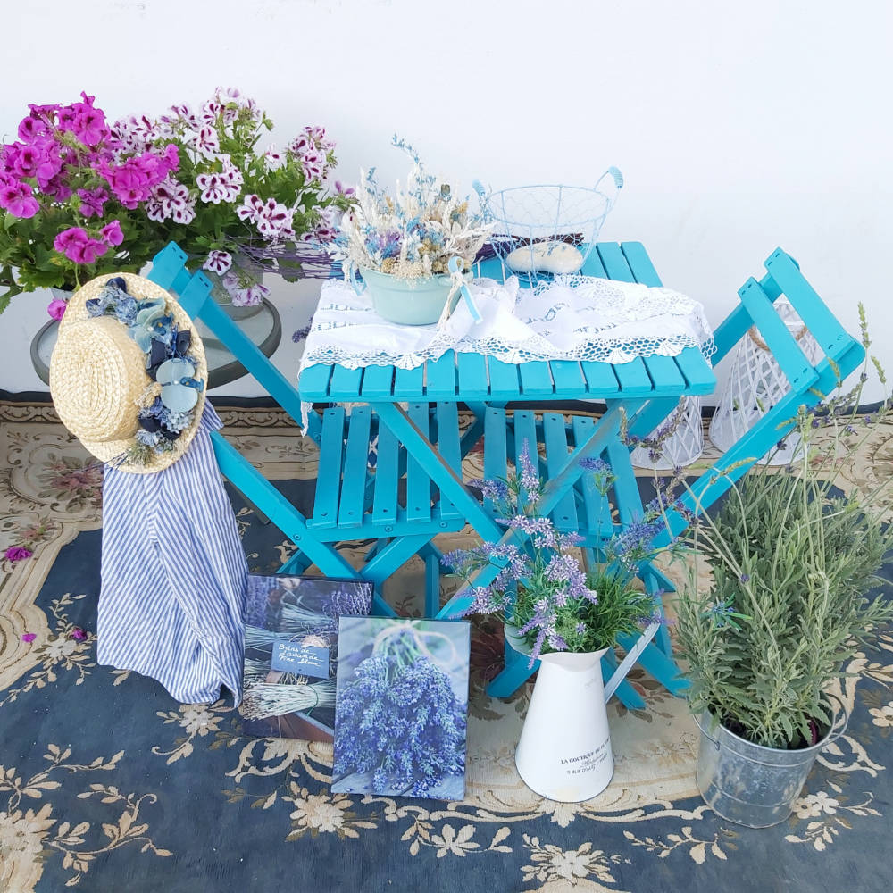 Una terrraza con muebles de madera de color azul, planta lavanda, flores preservadas, sombrero decorativo con flores y lienzos con fotografía de ramilletes de lavanda.