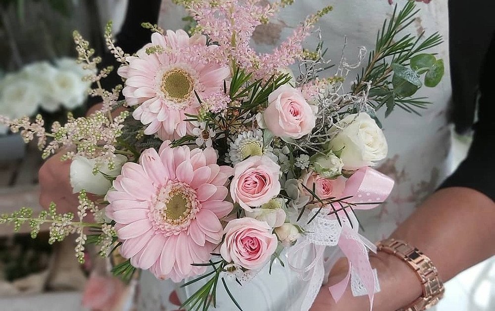 Centro de flores primaverales en tonos rosados con recipiente de cerámica blanco, sobre la mano de Nancy, equipo de las flores de Rita