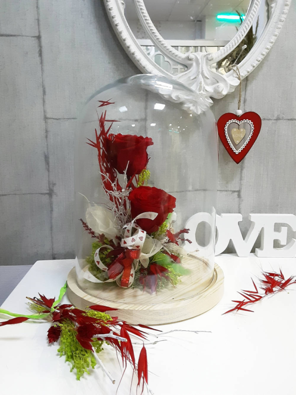 Arreglo con 2 rosas rojas preservadas y flores de secas de colores verdes y rojas en la base, en una cúpula de vidrio.